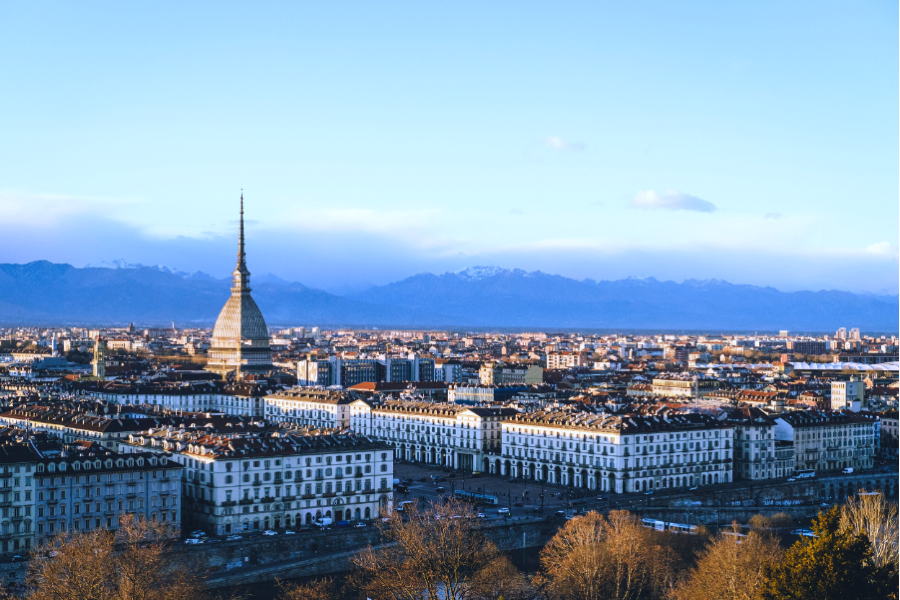 Panoramic view of a European city featuring a prominent domed tower, surrounded by historic buildings, with snow-capped mountains in the background.